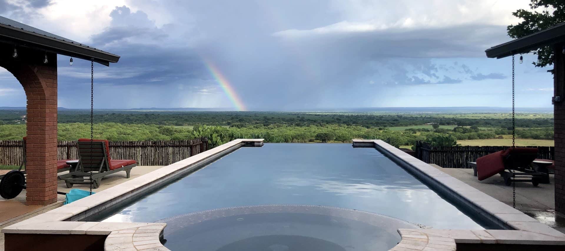 Large patio with infinity pool looking out in the distance with green trees, storm clouds, and rainbow