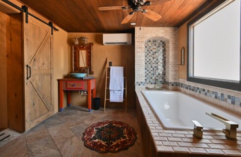 Large bathroom with light tan walls, tile flooring, bathtub surrounded by multicolored tile, multicolored vanity, and sliding barn door