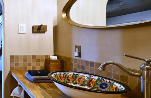 Close up view of a custom painted vanity sink on top of wooden counter with a multicolored tan tiled backsplash and large oval mirror on the wall