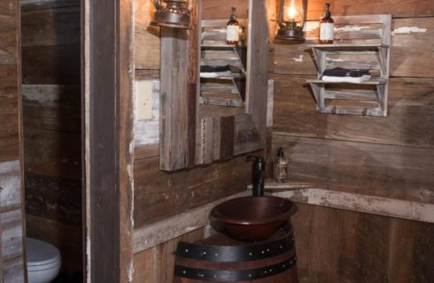 Corner of a bedroom that looks like a jail cell with copper bowl sink sitting on a whiskey barrel, wood framed mirror, and antiqued barn wood paneling on the walls