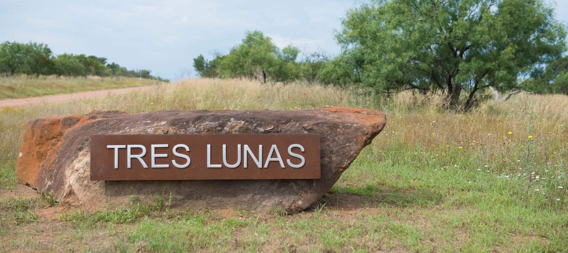 Large metal sign attached to large boulder that reads Tres Lunas surrounded by green grass and shrubs