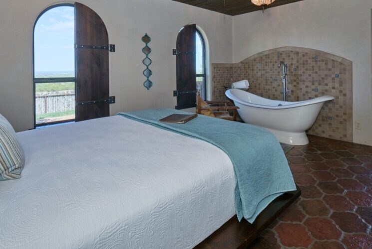 Bedroom with light tan walls, tiled flooring, white slipper tub, and bed with blue, white, green and tan bedding