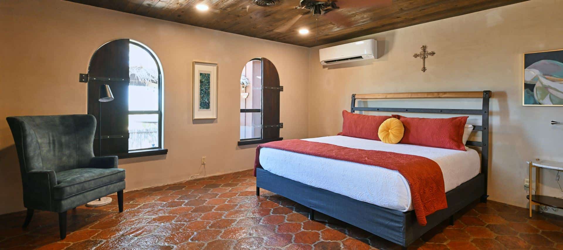 Bedroom with light colored walls, terracotta tile flooring, bed with white bedding, red pillows and blanket, and blue upholstered armchair