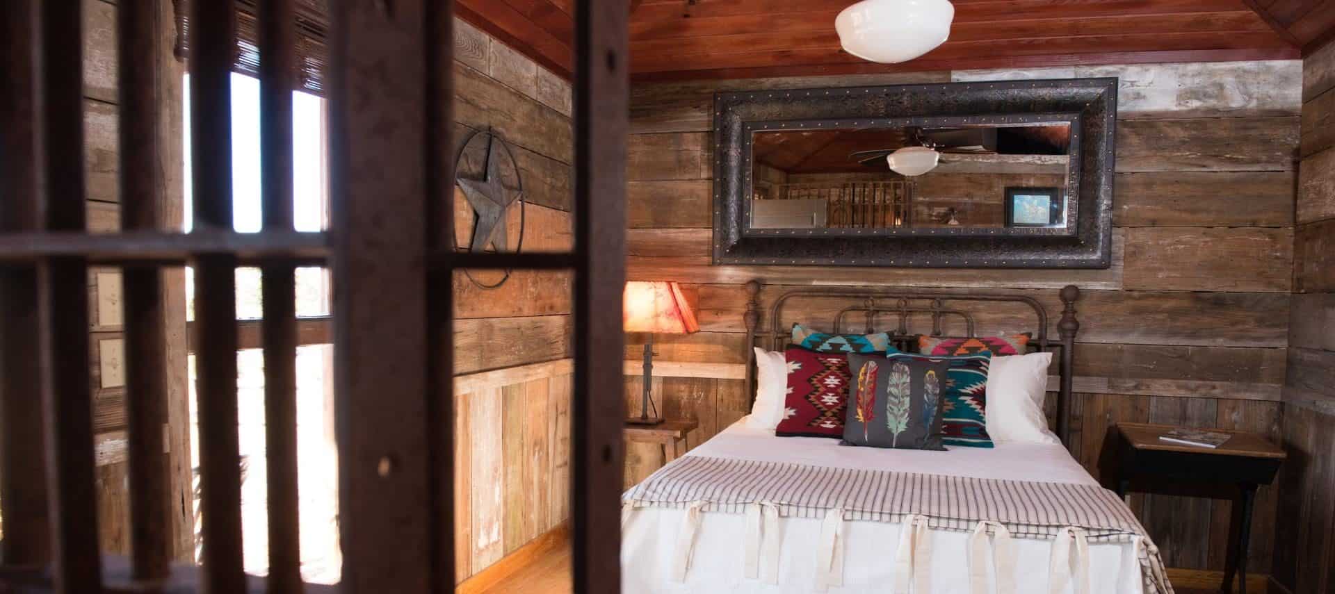 Bedroom that looks like a jail cell with antiqued barn wood paneling on the walls, hardwood flooring, wrought iron bed, and white bedding