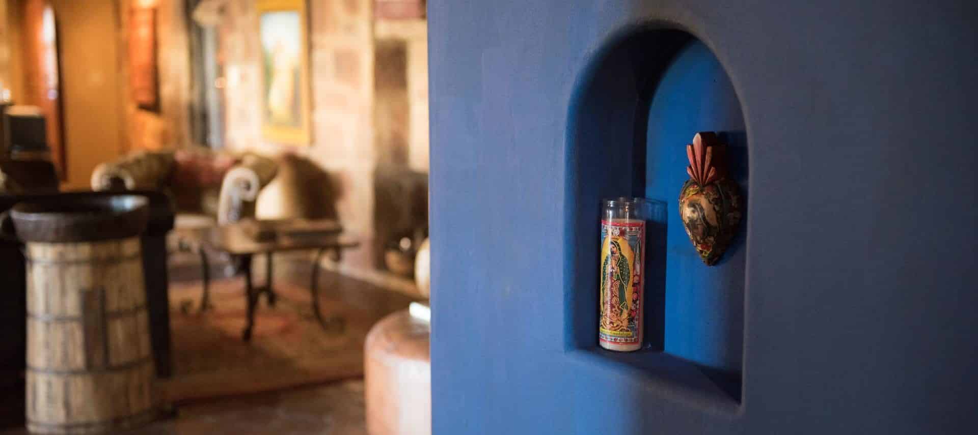 Close up view of blue wall with an arched recessed shelf, with a Catholic candle sitting inside and a hanging heart shaped piece of art