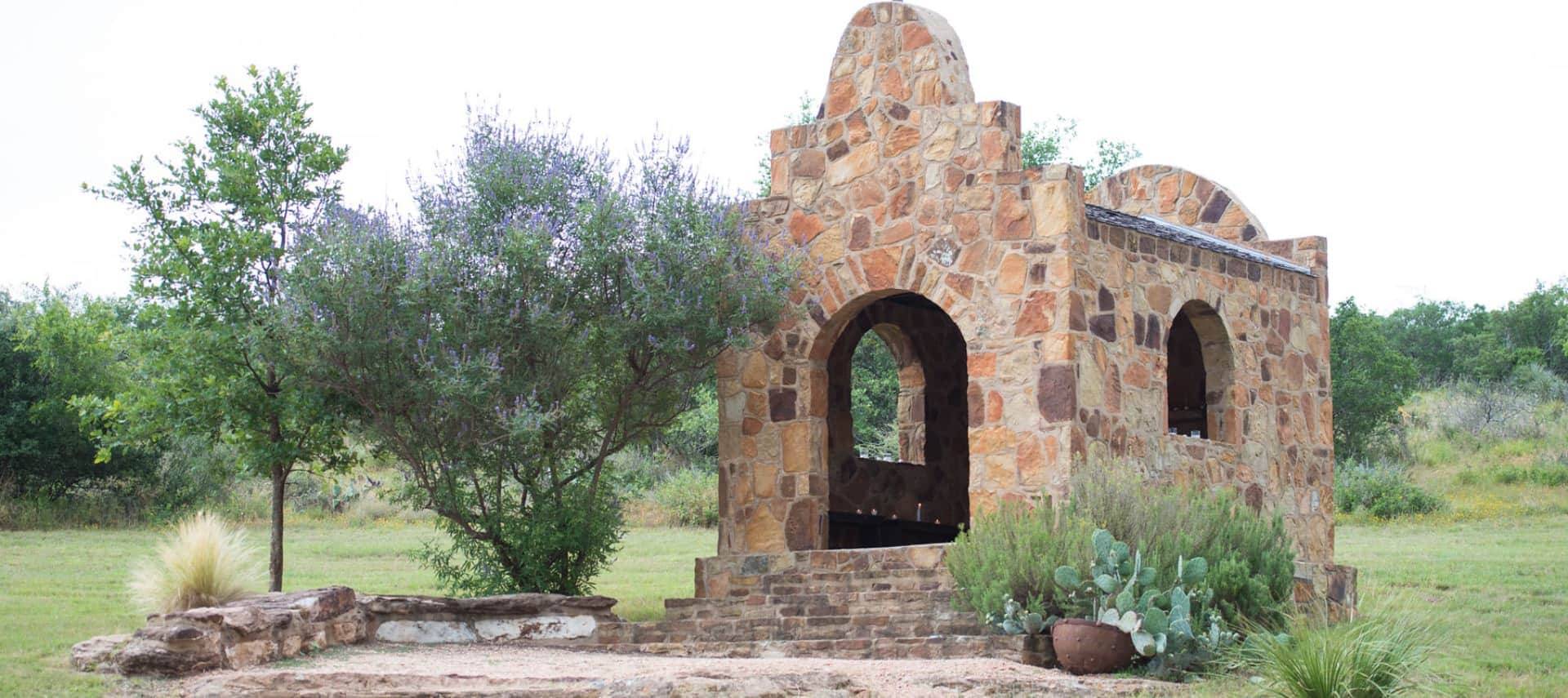 Old stone brick chapel surrounded by green grass, small trees and bushes, and cactus