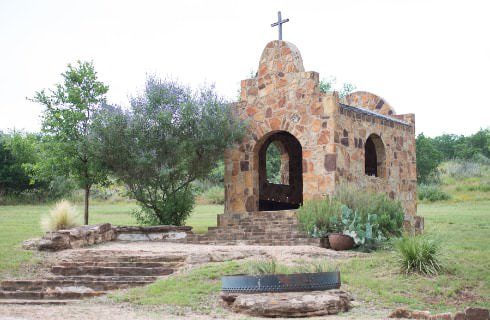 Old stone brick chapel surrounded by green grass, small trees and bushes, and cactus